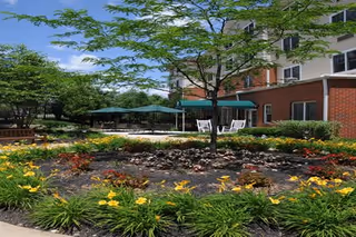 Landscaped outdoor courtyard with flower beds, trees, patio seating and a multi-story brick building in the background.