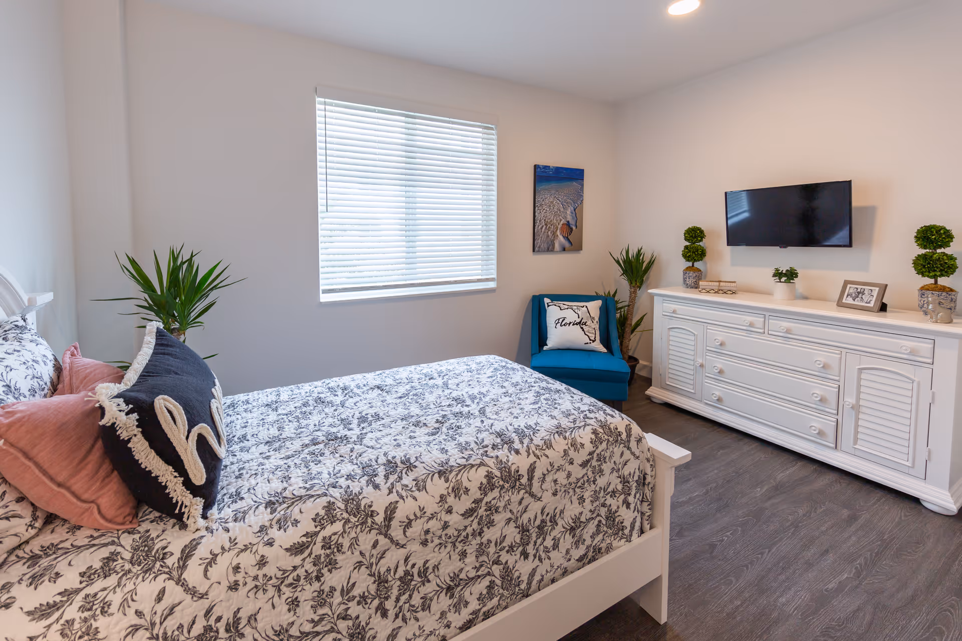 A bright bedroom featuring a bed with floral patterned bedding and decorative pillows, a window with blinds, a blue chair with a Florida-themed pillow, a white dresser with a flat-screen TV mounted above it, and several potted plants and framed photos on the dresser.