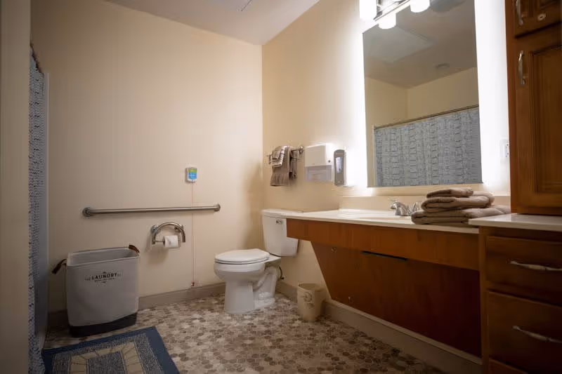 A clean bathroom with a toilet, a grab bar on the wall, a laundry basket, a large mirror with lights, a sink with folded towels on the counter, and a wooden cabinet.