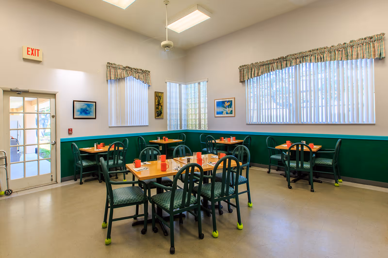A dining room in an assisted living facility with several wooden tables and green cushioned chairs. Each table is set with red mugs, napkins, and utensils. The room has large windows with vertical blinds and valances, light-colored walls with a green wainscot, and framed artwork on the walls. There is an exit door with a glass panel and an exit sign above it.