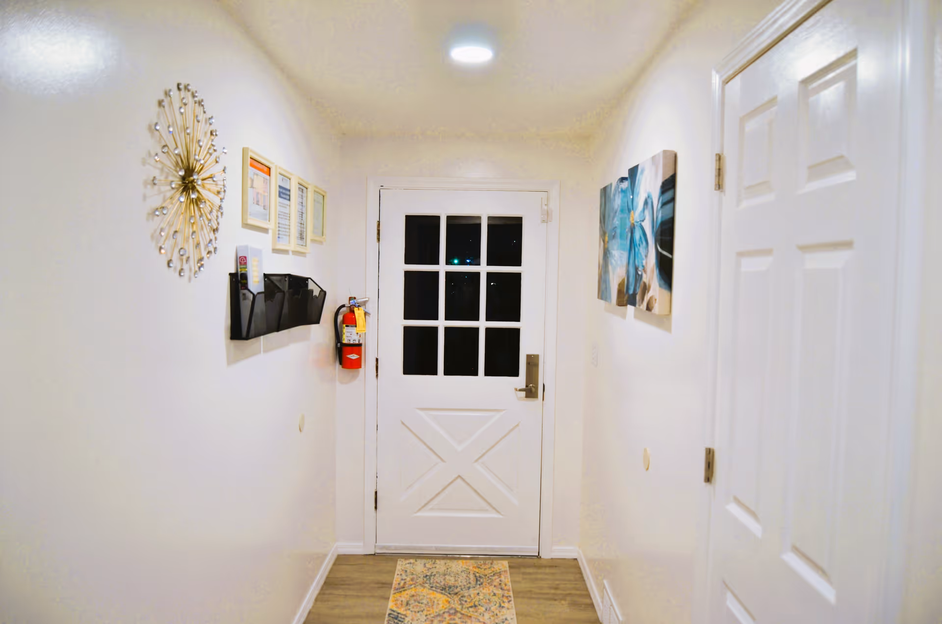 A narrow hallway with white walls and wooden flooring leading to a white door with glass panes. On the left wall, there is a decorative sunburst wall clock, three framed documents, a black wall-mounted organizer, and a fire extinguisher. On the right wall, there are two blue floral paintings and a closed white door. A small patterned rug is placed on the floor in front of the door.