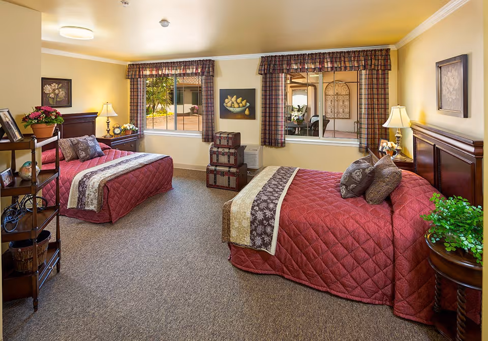A cozy bedroom with two beds covered in red quilted bedspreads and decorative pillows. The room has beige walls, carpeted floor, two windows with plaid valances, and framed artwork on the walls. There are bedside tables with lamps and plants, a small shelving unit with decorative items, and a stack of three vintage-style suitcases near the window.