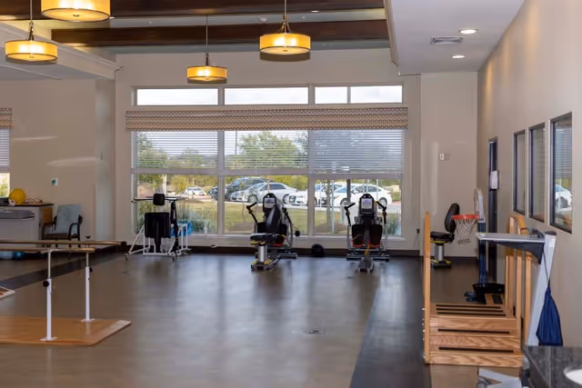Spacious interior therapy/fitness room with exercise equipment, parallel bars, and large windows looking out to a parking area.