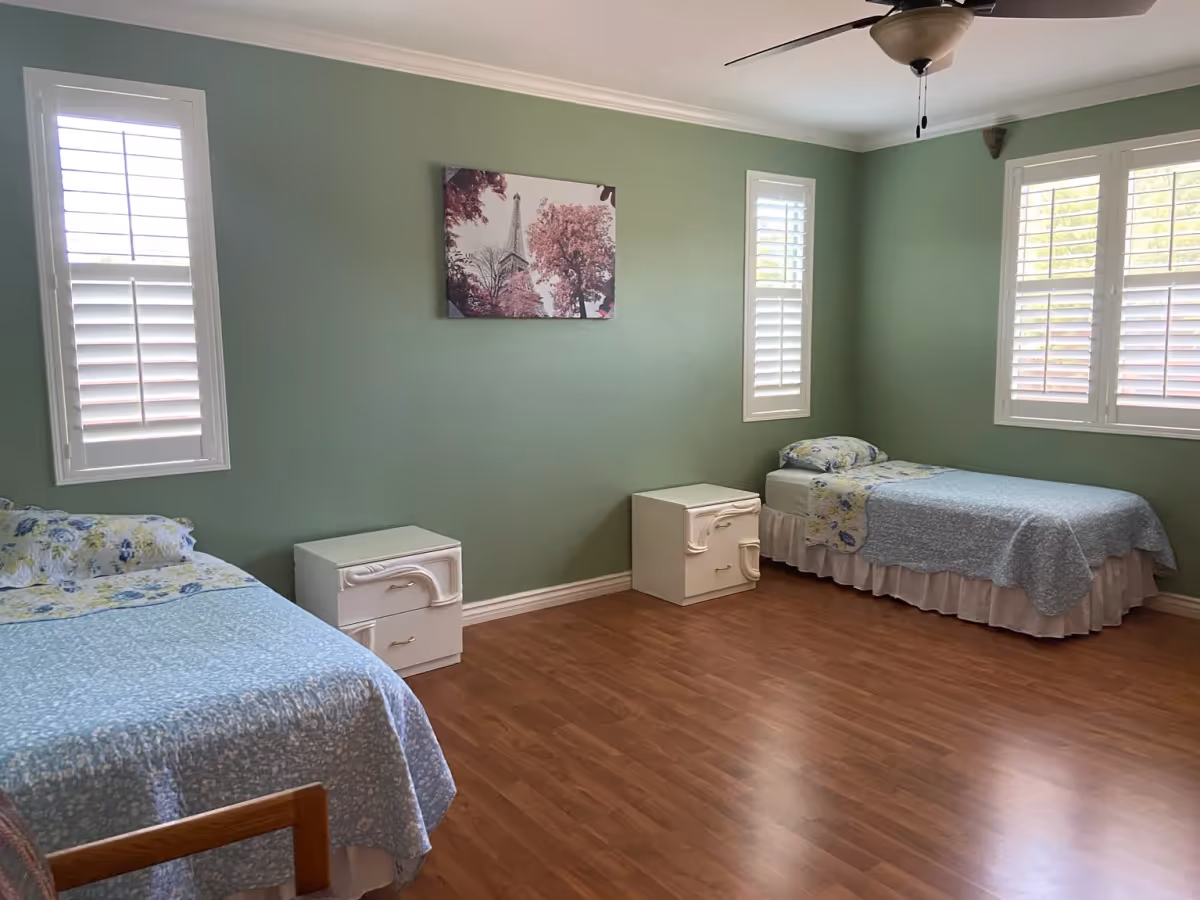 A bedroom with two single beds placed against light green walls. Each bed has a floral pillow and a blue bedspread. Between the beds are two white nightstands with drawers. The room has three windows with white plantation shutters allowing natural light to enter. A ceiling fan with a light fixture is mounted on the white ceiling. A framed picture of the Eiffel Tower surrounded by pink trees hangs on the wall between the windows.