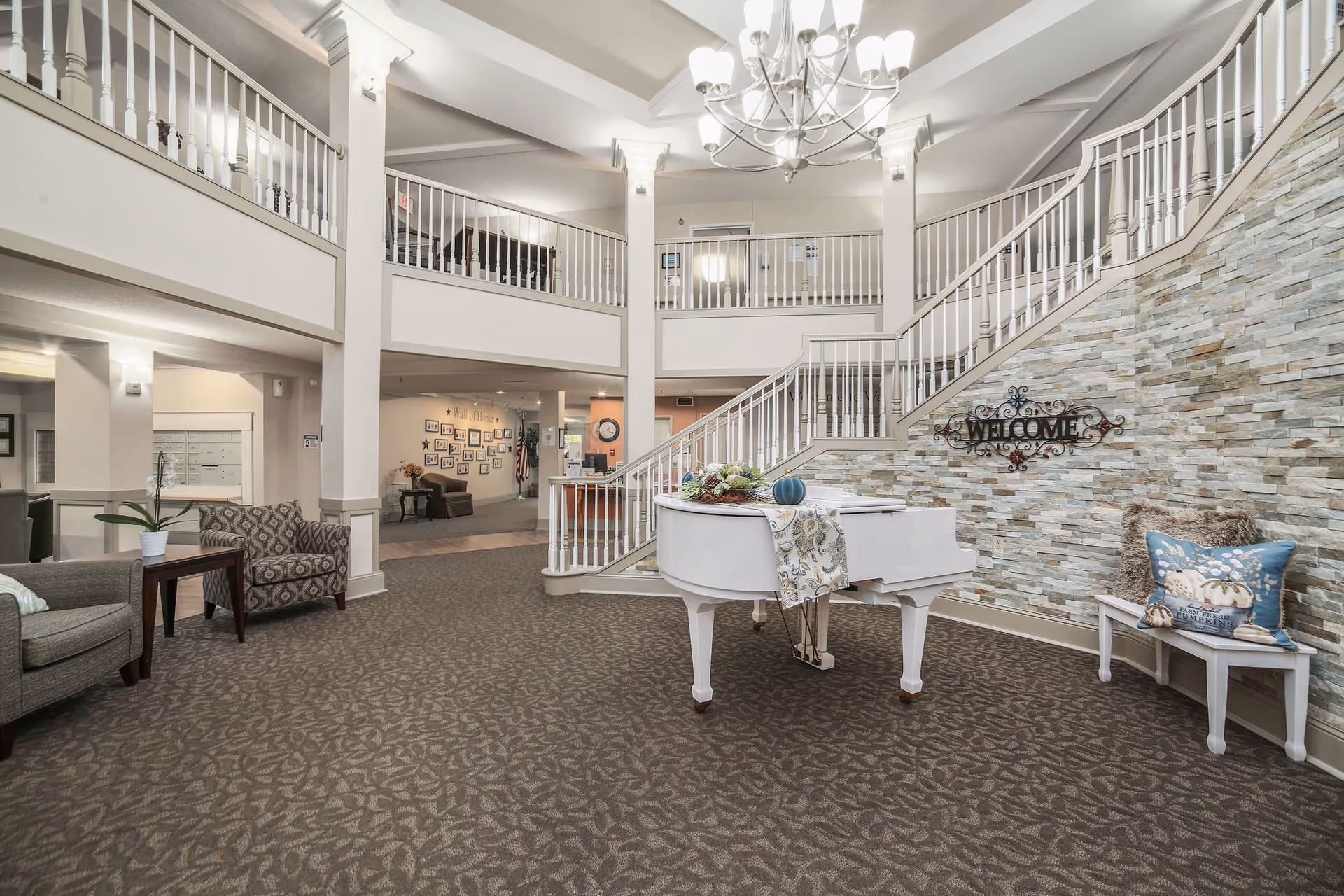 Spacious senior living facility lobby with a white grand piano decorated with a table runner and floral arrangement, a stone accent wall with a decorative 'WELCOME' sign, a staircase with white railings leading to an upper balcony, patterned carpet flooring, and seating areas with chairs and a small table.