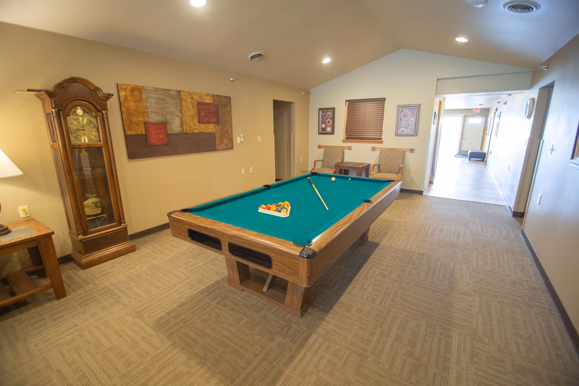 Interior room with a pool table in the center, a grandfather clock to the left, two chairs with a small table against the far wall, and a hallway leading to an exit door in the background.