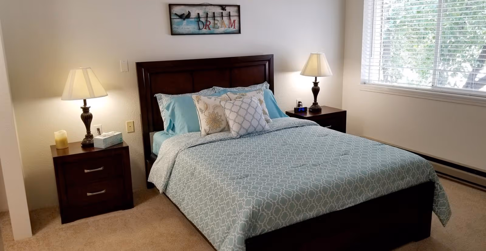 Neatly made bedroom with a bed, two nightstands and lamps, a window, and wall art above the headboard.