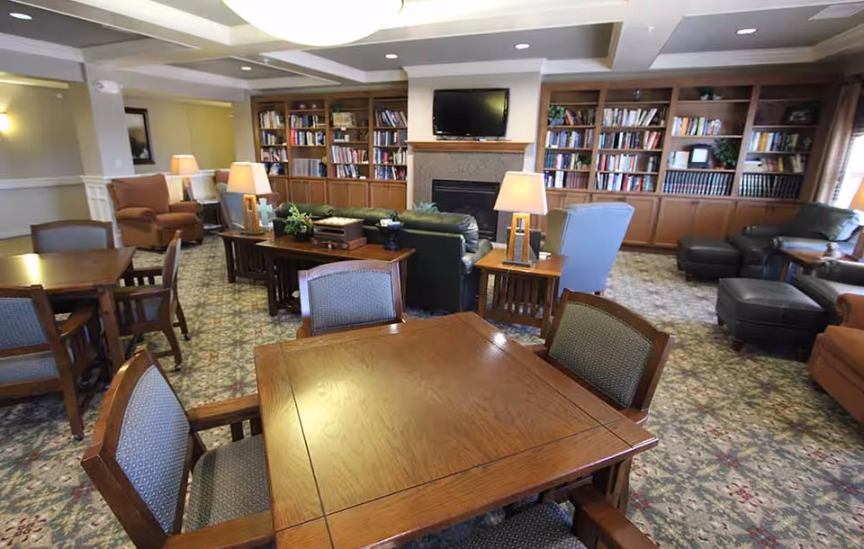 Communal lounge with wooden tables and chairs, upholstered seating, lamps, built-in bookshelves, a fireplace and wall-mounted TV.