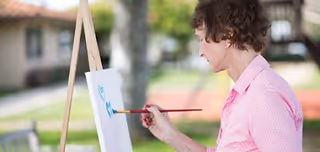 An elderly woman wearing a pink shirt painting on a canvas set on an easel outdoors, with a blurred background of a building and greenery.