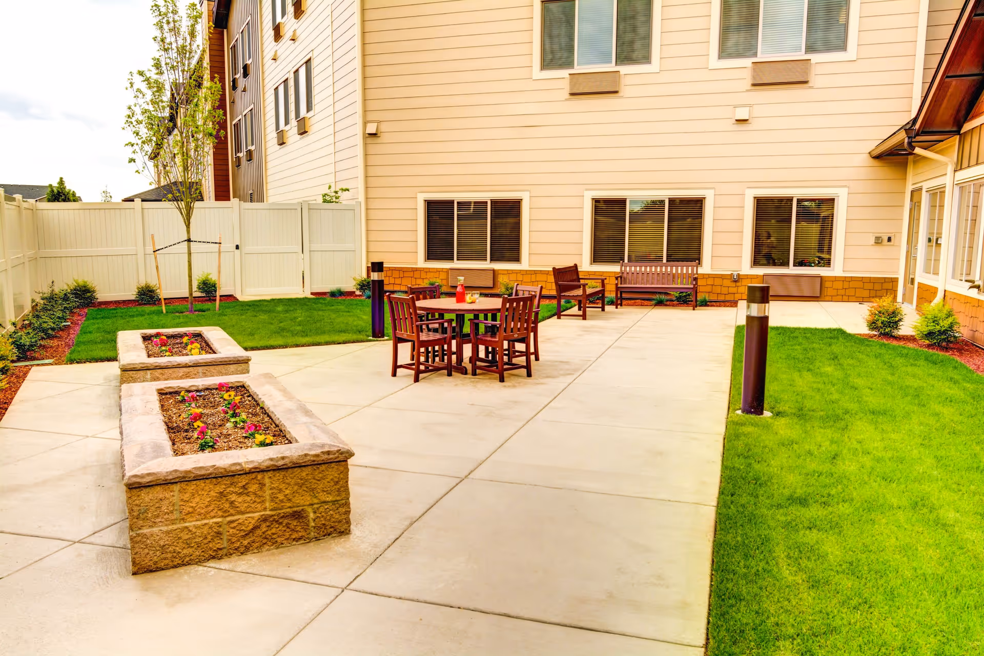Outdoor patio area at Village at Keizer Ridge with concrete walkways, two rectangular raised flower beds with colorful flowers, green grass lawns, a young tree, wooden table with four chairs, and wooden benches against the building wall.