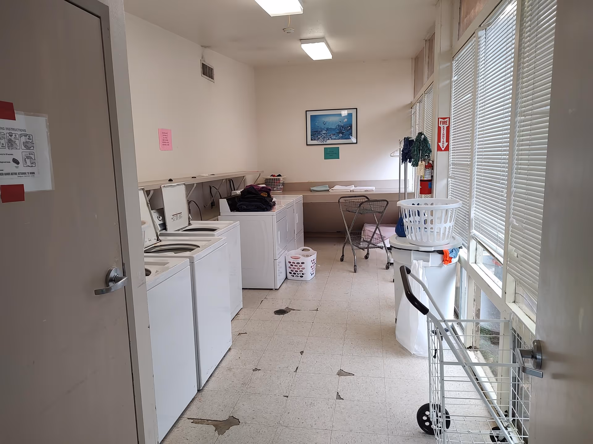 Laundry room with multiple washing machines and dryers along the left wall, laundry baskets on top and on the floor, a laundry cart near the door, and large windows with blinds on the right side. The floor has some worn and damaged tiles, and there is a fire extinguisher mounted on the right wall.
