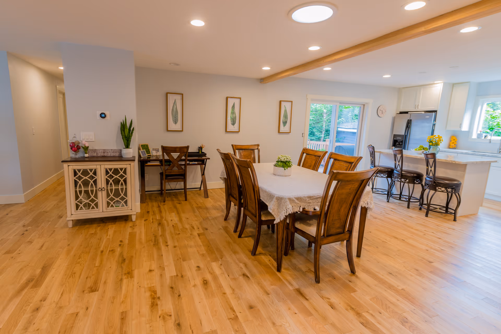 A bright, open dining area with a wooden dining table covered with a lace tablecloth and surrounded by eight wooden chairs. To the right is a kitchen island with three high chairs, a refrigerator, and white cabinetry. The room has wooden flooring, recessed lighting, and three framed leaf prints on the wall. A sliding glass door leads outside, allowing natural light to fill the space.