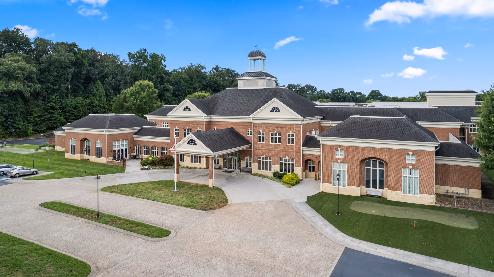 Front exterior view of a large brick building with multiple sections and a central entrance covered by a portico. The building has a cupola on the roof and is surrounded by a paved driveway and green lawns with trees in the background under a partly cloudy blue sky.