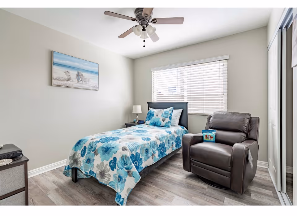 A bright bedroom with a single bed covered in a blue and gray floral quilt, a matching pillow, a dark brown leather recliner chair with a small colorful pillow on it, a nightstand with a lamp and alarm clock, a ceiling fan with lights, a large window with white blinds, and a beach-themed painting on the wall.