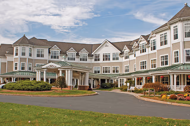 Front exterior of a large multi-story senior living building with a covered entrance, driveway, and landscaped grounds.