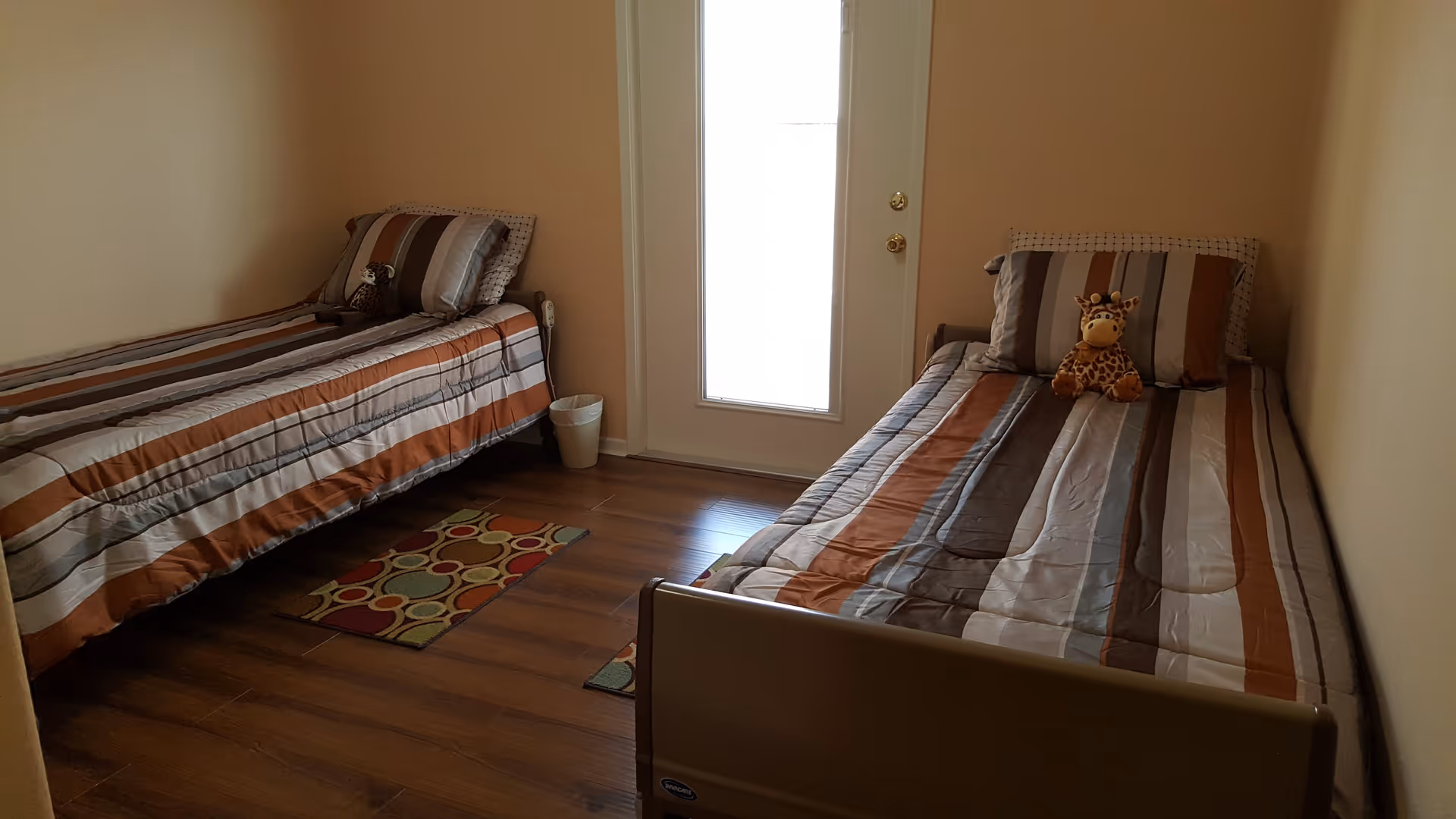 A small bedroom with two single beds placed against opposite walls. Each bed has striped bedding in shades of brown, gray, and white, with a pillow and a stuffed giraffe toy on each bed. The room has wooden flooring, two small colorful rugs, beige walls, and a door with a frosted glass panel letting in light.