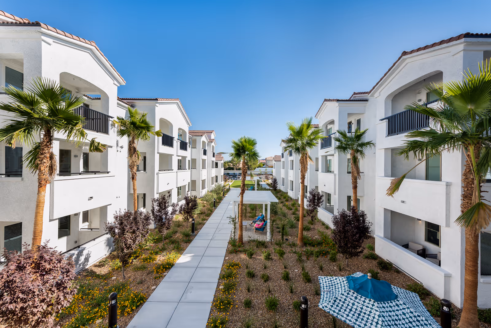 View of a modern senior living facility courtyard with white multi-story buildings on both sides, palm trees, landscaped garden beds, a paved walkway, and a shaded seating area with two people sitting under it. A blue and white patterned umbrella is visible in the foreground.