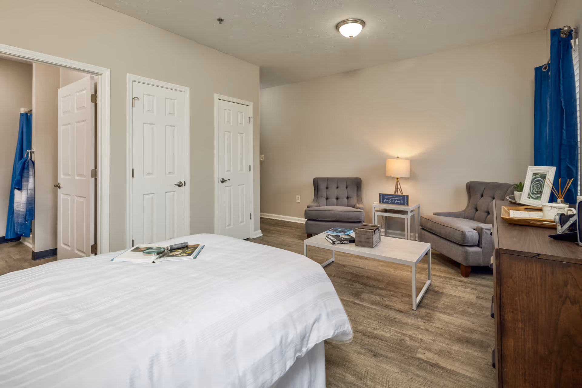 A bedroom with a white bedspread in the foreground, two gray upholstered chairs and a matching loveseat around a white coffee table with books and a decorative box. A small side table with a lamp is against the wall, and a wooden dresser with framed pictures and decor is on the right. Three white doors are visible on the left wall, one leading to a bathroom with a blue towel hanging inside. The room has wood flooring and neutral-colored walls with a blue curtain on the right window.