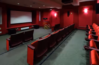 Interior view of a small movie theater or screening room with red walls and carpet, rows of red cushioned seats with cup holders, a large white projection screen at the front, and a vintage popcorn machine near the screen.