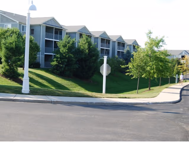 Exterior view of a multi-story residential building with balconies, surrounded by green trees and a well-maintained lawn. A sidewalk runs alongside the building, and a person is walking in the distance. There are street lamps and a stop sign near the road in the foreground.
