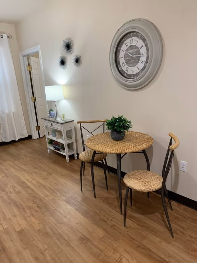 A small dining area with a round wicker table and two matching chairs on a wooden floor. A potted green plant is centered on the table. On the wall above the table is a large round clock with Roman numerals. To the left, there is a white side table with a lamp, some books, and small decorative items. The room has light-colored walls and a door partially open leading to another room.