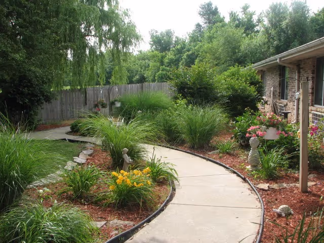A curved concrete walkway bordered by lush green plants, flowers, and mulch leading alongside a brick building with a covered porch. Trees and a wooden fence are visible in the background under a clear sky.