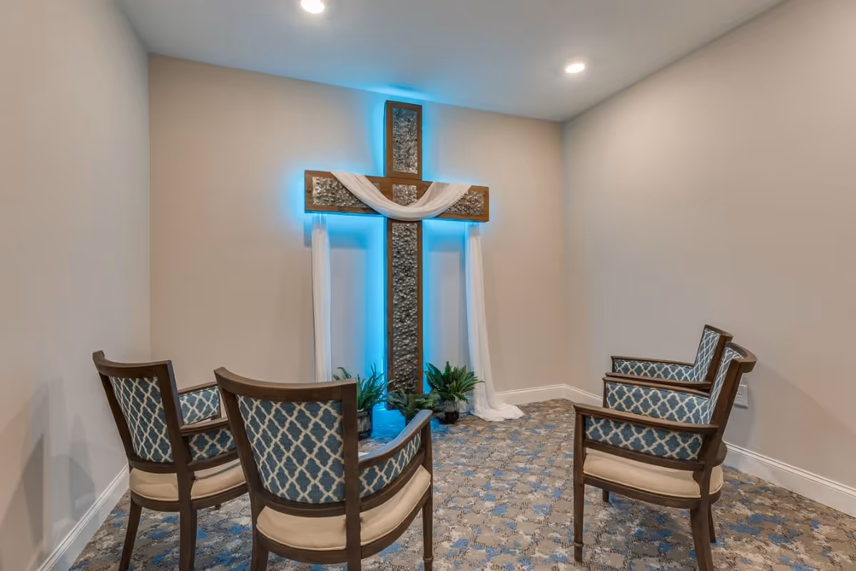 A small room with four wooden chairs upholstered in blue patterned fabric arranged in a semicircle facing a large decorative cross on the wall. The cross is backlit with blue lighting and draped with white fabric, with some green plants at its base. The room has beige walls and a patterned carpet with blue and beige tones.
