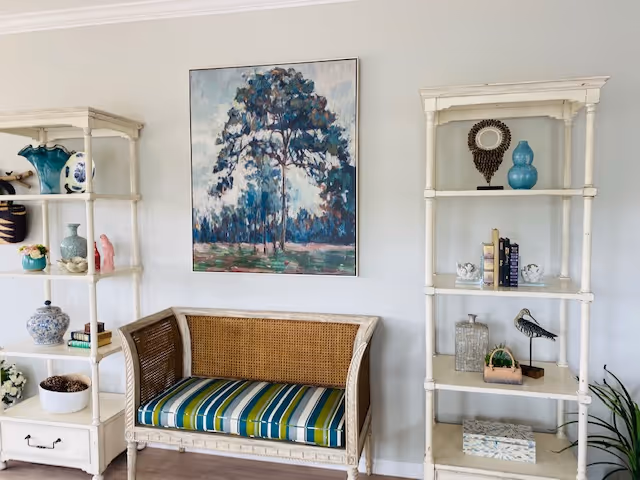 A cozy seating area with a wicker loveseat featuring a striped blue, green, and white cushion. On either side of the loveseat are white wooden shelving units displaying decorative items such as vases, books, and small sculptures. A large painting of a tree with a blue and green color palette hangs on the light gray wall behind the loveseat.