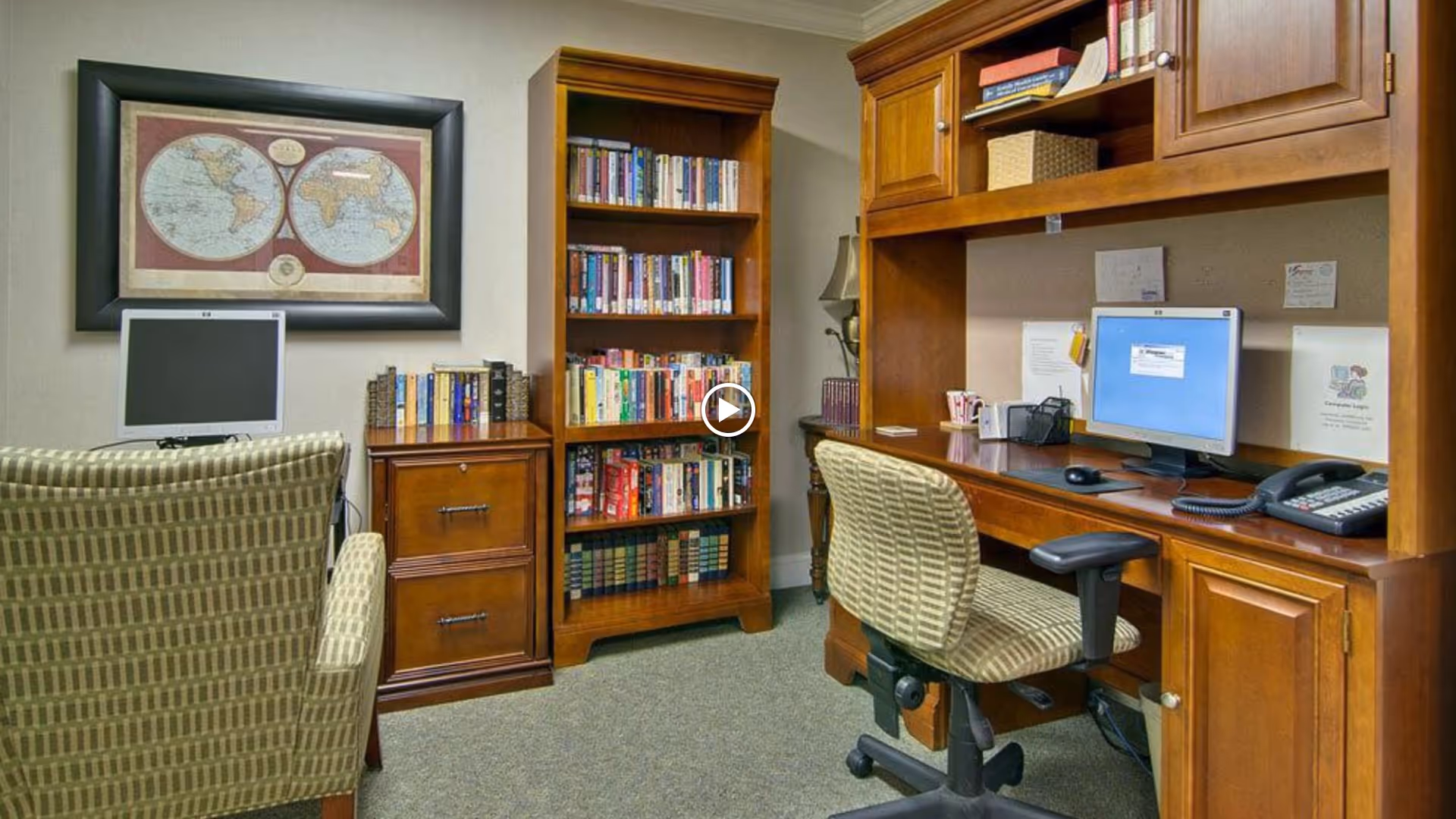 A cozy office or study area in a senior living facility featuring two computer workstations with monitors, a wooden bookshelf filled with books, a wooden filing cabinet, and comfortable patterned office chairs. A framed world map hangs on the wall above one workstation.