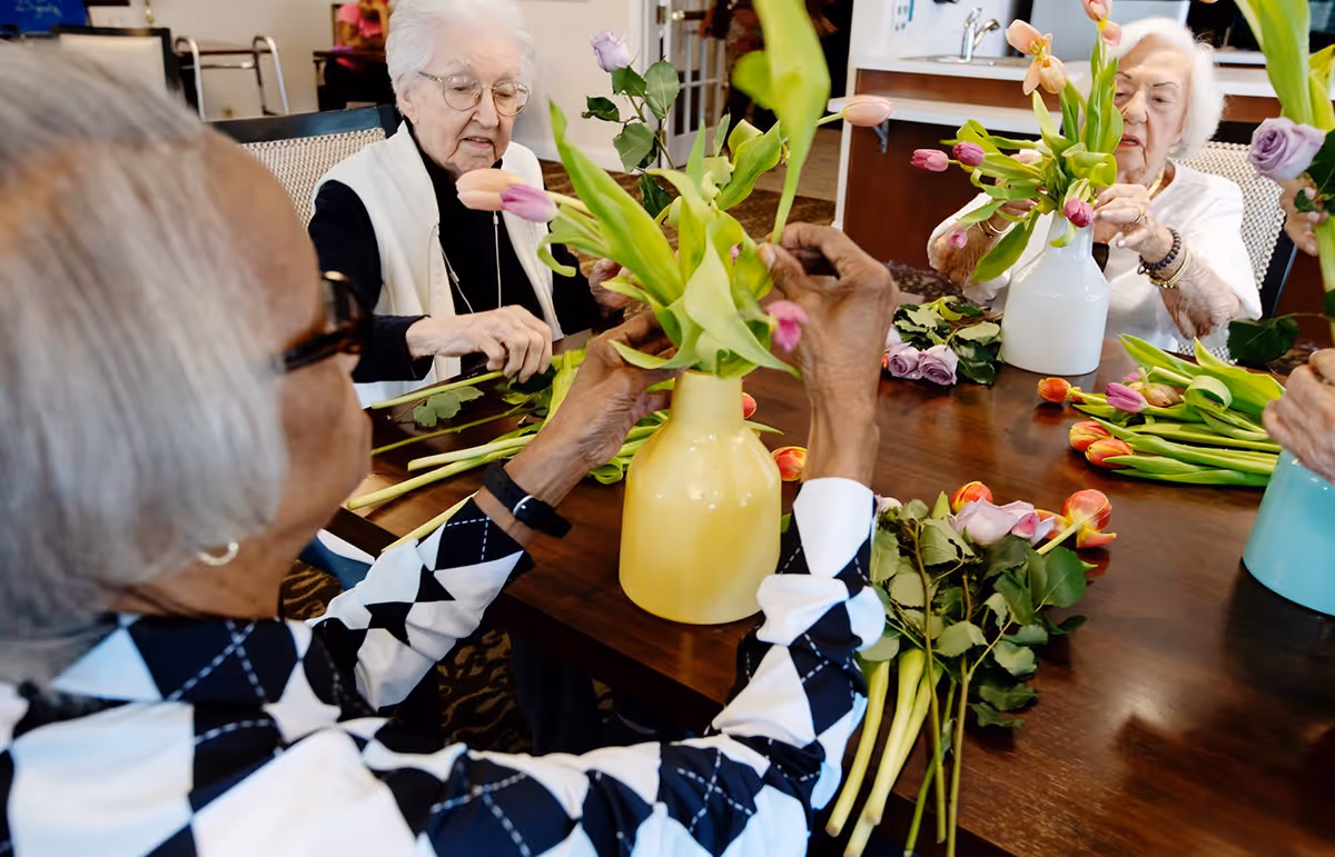 A group of elderly women sitting around a wooden table arranging colorful tulip flowers in vases inside a well-lit room.