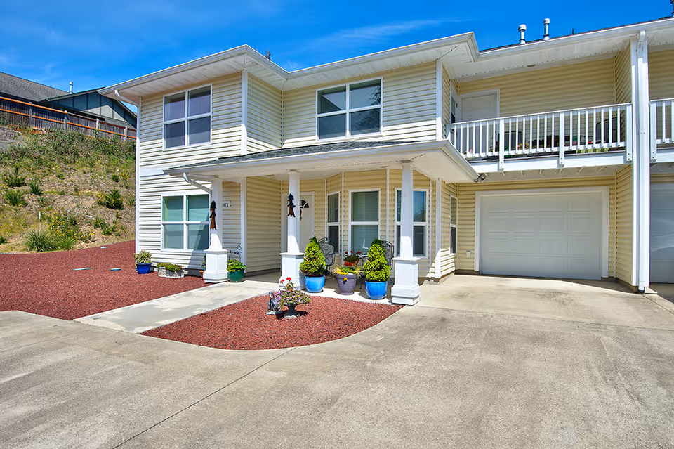 Exterior view of a two-story residential building with beige siding, multiple windows, a covered porch with white columns, potted plants, and a concrete driveway leading to a garage. The area around the porch is landscaped with red mulch and small shrubs.
