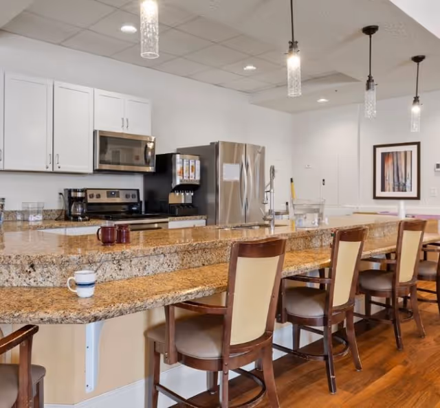 A modern kitchen area with a long granite countertop and several wooden chairs with cushioned seats. The kitchen features white cabinets, a stainless steel microwave, stove, refrigerator, and a beverage dispenser. Pendant lights hang from the ceiling, and there is a framed artwork on the wall in the background.