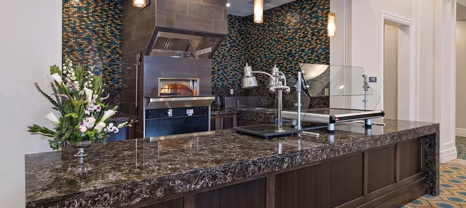 A modern kitchen area with a dark marble countertop and a built-in pizza oven with a stainless steel hood. The backsplash features a mosaic tile design in shades of blue, brown, and beige. There is a glass sneeze guard on the counter, two heat lamps, and a floral arrangement on the left side. The floor has a patterned carpet, and there are white walls and doorways in the background.
