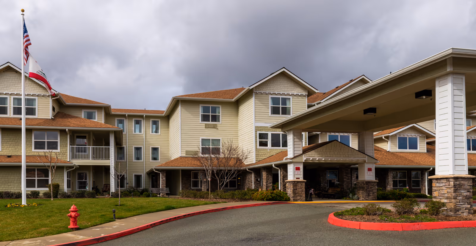 Front entrance of a three-story beige senior living building with a covered drop-off, flagpole, and red curb under a cloudy sky.