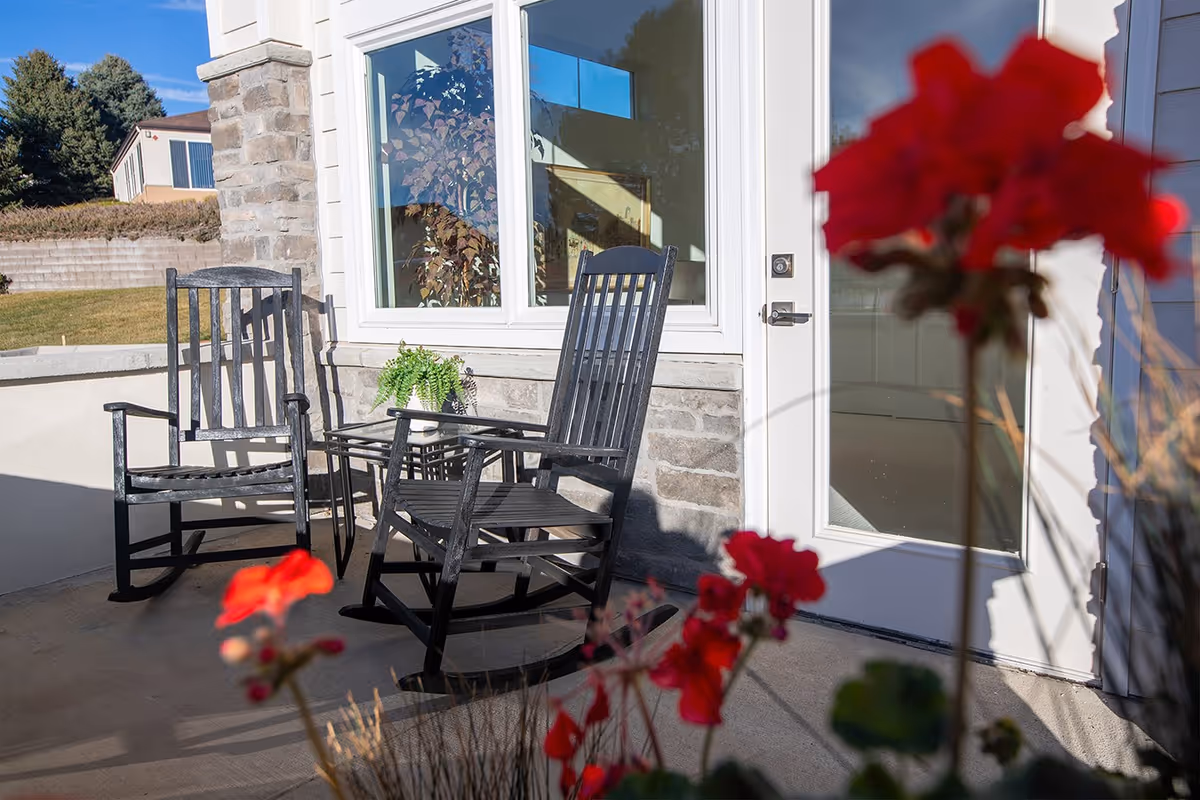 Outdoor patio area with two black rocking chairs and a small glass table with a potted plant, set against a stone and white wall exterior with a window and a glass door. Red flowers are visible in the foreground.