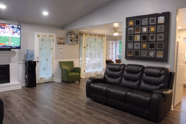 A modern living room with a black leather sofa, a green armchair, and a wall-mounted flat-screen TV showing a sports game. The room has wood flooring, light gray walls, recessed ceiling lights, and a decorative square wall art piece. There is a water dispenser near a glass door with sheer curtains, and an adjoining dining area is visible through an open space.