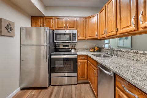 A kitchen with wooden cabinets, granite countertops, and stainless steel appliances including a refrigerator, stove with oven, microwave, and dishwasher. There is a sink with a faucet and a decorative bowl of fruit on the counter.