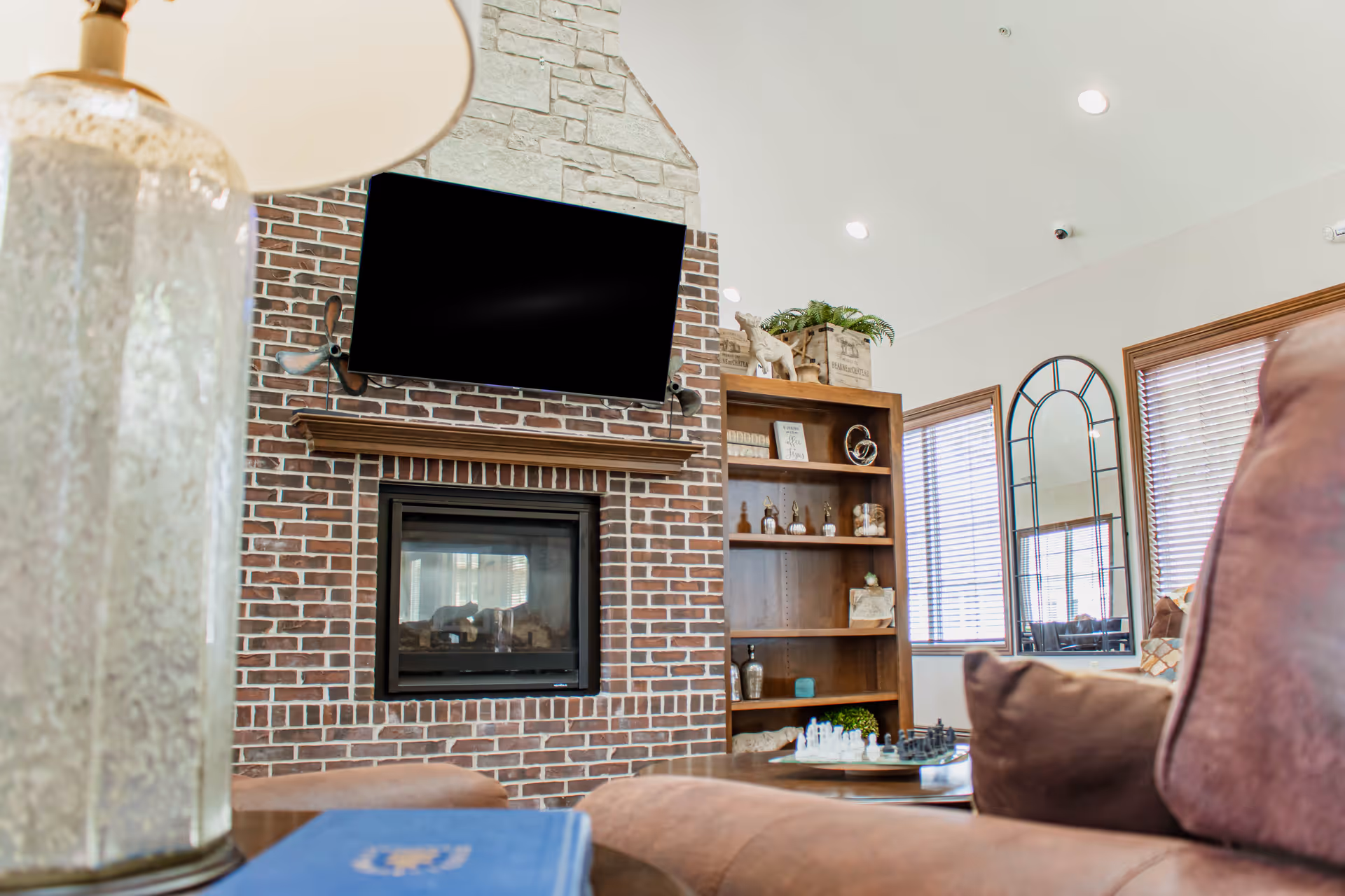 Cozy living room area with a brick fireplace and a mounted flat-screen TV above it. A wooden bookshelf with decorative items is next to the fireplace. There are windows with blinds and a large arched mirror on the wall. In the foreground, there is a brown cushioned chair and a table with a chess set and a lamp.