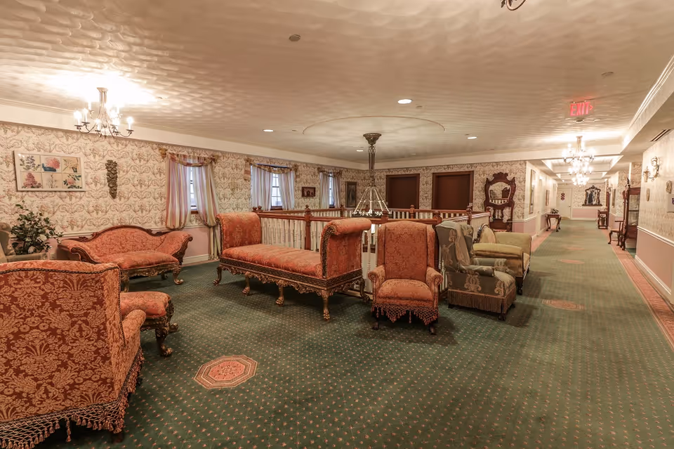 A spacious, vintage-style sitting area with ornate upholstered chairs and sofas arranged on a green carpet with patterned accents. The walls are decorated with floral wallpaper and framed artwork, and the ceiling features recessed lighting and a chandelier. A hallway extends to the right with additional antique furniture and wall sconces.