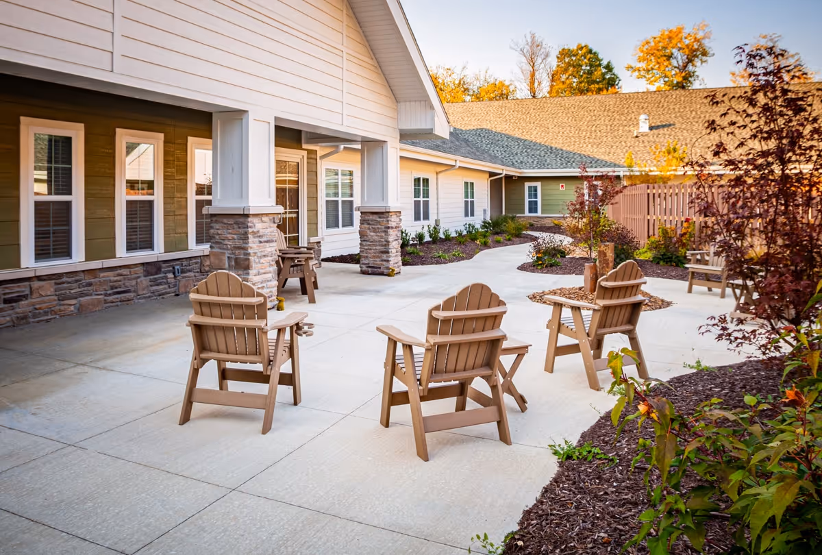 Outdoor patio area at Ciel of Fayetteville with several wooden Adirondack chairs arranged around a fire pit, surrounded by landscaped garden beds and a building with multiple windows and stone pillars.