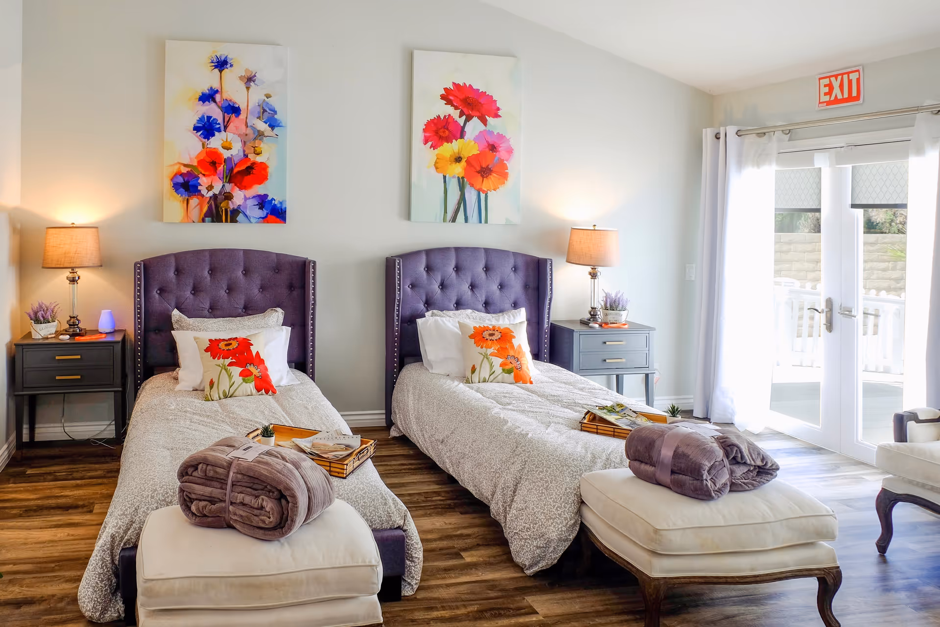 A bright bedroom with two twin beds featuring purple tufted headboards, floral pillows, and light gray bedding. Each bed has a folded blanket on a cushioned bench at the foot. There are two nightstands with lamps and small plants, colorful floral paintings above the beds, and a glass door with white curtains leading outside. An exit sign is visible above the door.