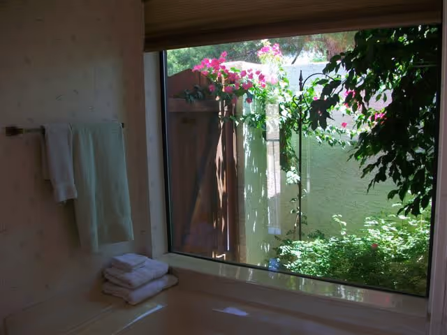 Bathtub with folded towels on the ledge beside a large window looking out onto a small garden with flowering vines.