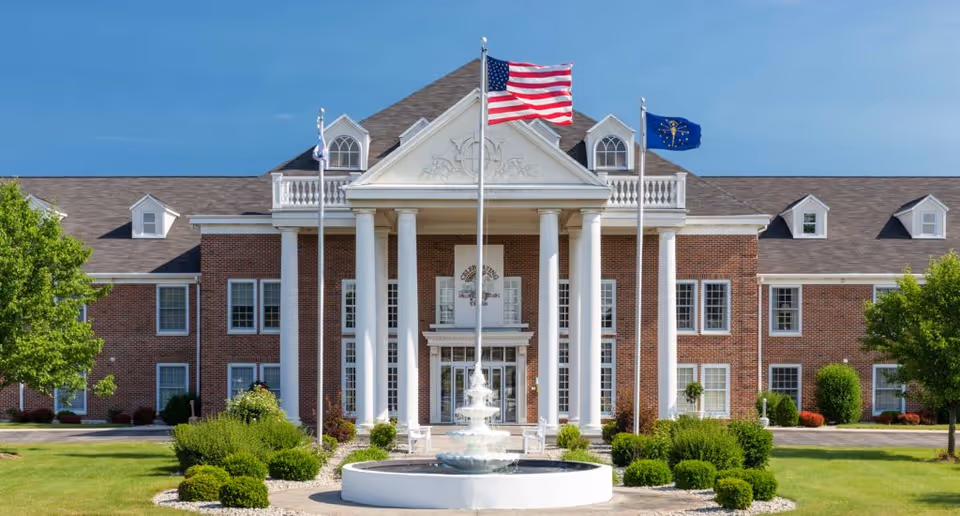 Front exterior view of The Towne House Retirement Community building with large white columns, a fountain in the foreground, and three flagpoles displaying the American flag and other flags, surrounded by green bushes and trees under a clear blue sky.