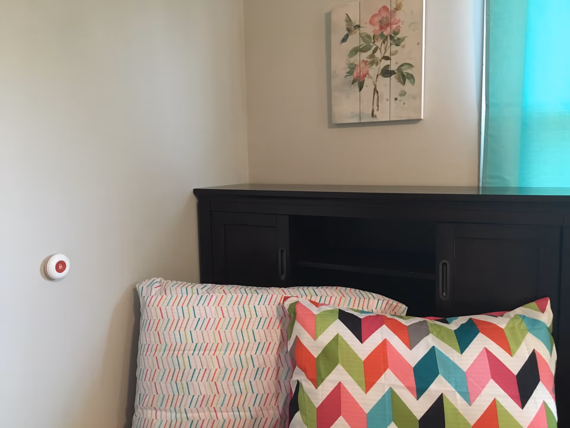 A corner of a bedroom with a dark wooden headboard, two colorful patterned pillows, a wall-mounted emergency call button, a window with a teal curtain, and a floral painting with a hummingbird above the headboard.
