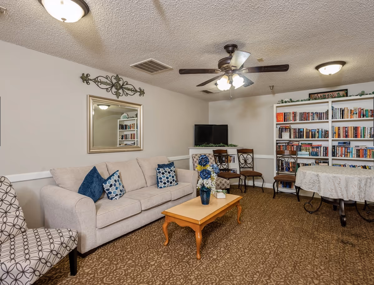 A cozy living room area with a beige sofa adorned with blue and patterned throw pillows, a wooden coffee table with a vase of blue and white flowers, patterned armchairs, a ceiling fan with lights, a TV on a white stand, and a large bookshelf filled with books. There is also a round table covered with a white tablecloth and several chairs around it.