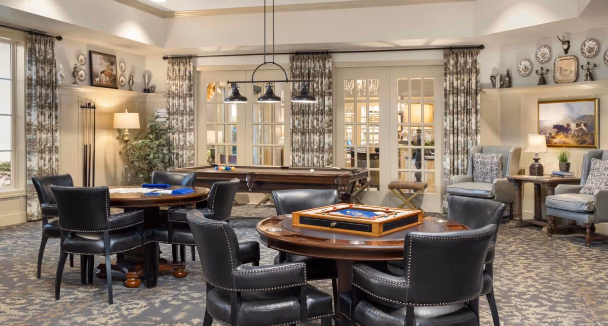 A well-lit, cozy common area with multiple round wooden tables surrounded by black leather chairs. One table has a backgammon game set up. In the background, there is a pool table and French doors with glass panes. The room features patterned curtains, a carpet with a floral design, wall decorations including plates and framed artwork, and several lamps providing warm lighting.