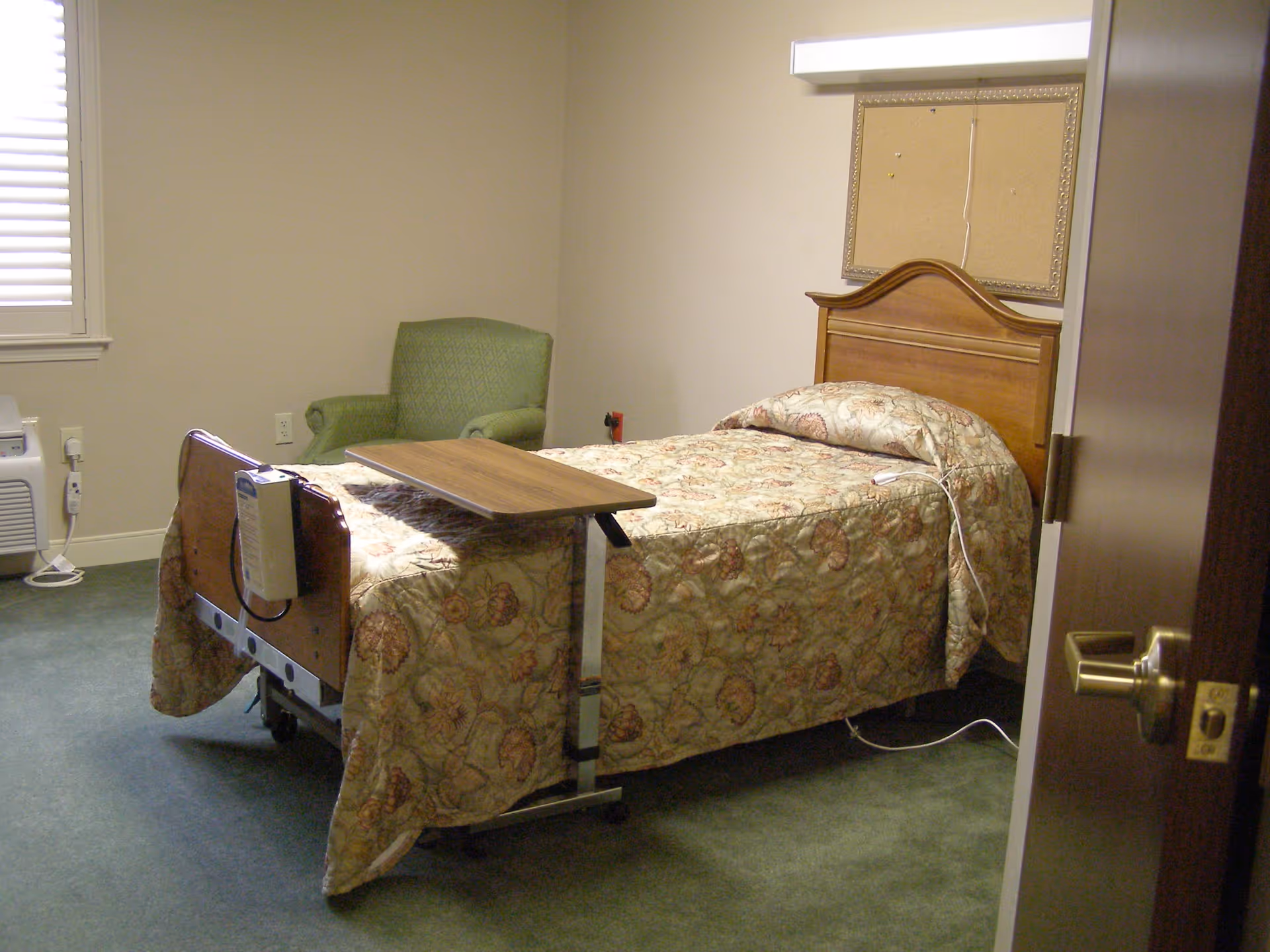 A small nursing facility bedroom with a single bed covered in a floral patterned quilt. The bed has a wooden headboard and a movable overbed table positioned on top. There is a green upholstered armchair in the corner near a window with closed blinds. The room has beige walls, green carpet, and a cork bulletin board mounted on the wall above the bed.