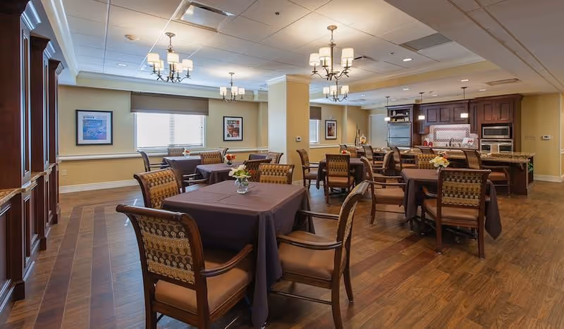 Communal dining room with multiple tables covered in dark tablecloths, upholstered chairs, chandeliers, and an open kitchen area in the background.