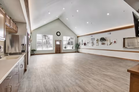 A spacious, empty room with high vaulted ceilings and recessed lighting. The room features large windows letting in natural light, a wall clock above a door, and a long wall decorated with various framed pictures and plants. On the left side, there is a kitchenette area with cabinets, a sink, and a stainless steel refrigerator. The floor is covered with light wood-style flooring.