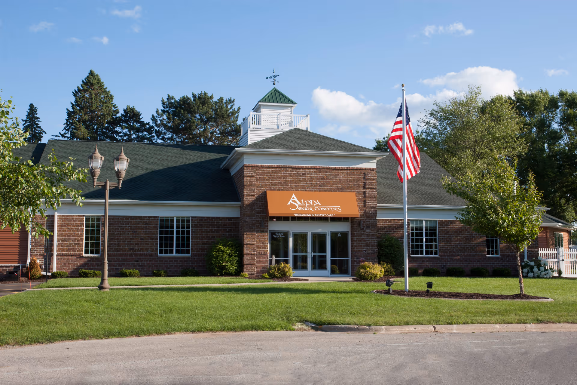 Exterior front view of Alpha Senior Concepts - Suamico Property, a single-story brick building with a green roof, an American flag on a flagpole, two street lamps, and well-maintained lawn and trees under a blue sky with some clouds.